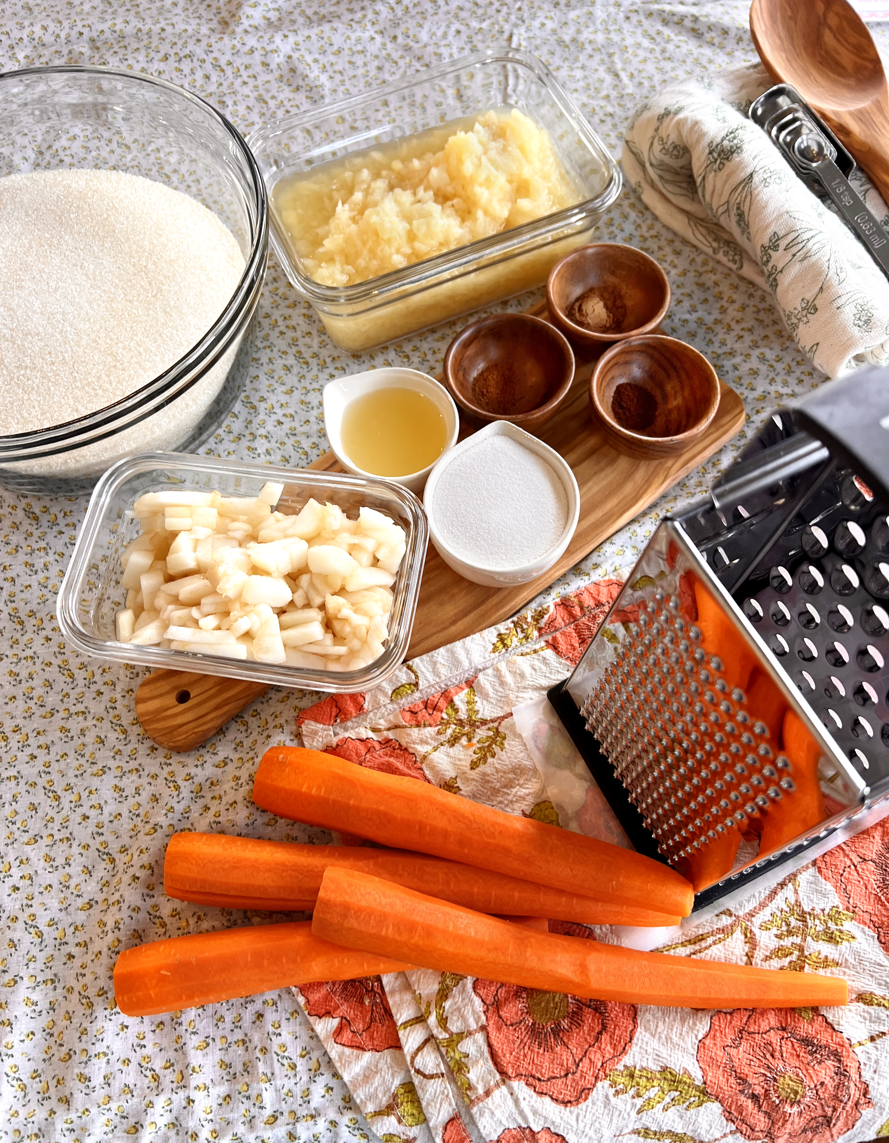 Ingredients laid out for carrot cake jam including fresh carrots, pineapples, sugar, spices, and a grater and measuring bowls on a rustic surface